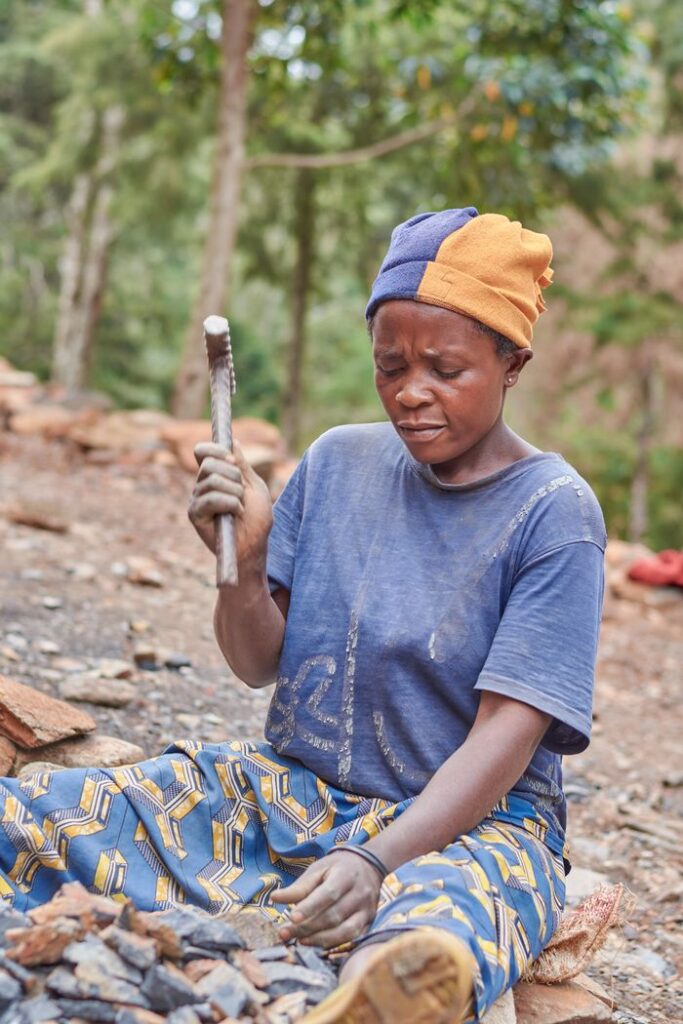 An African woman wearing a colorful headwrap and patterned pants is working outdoors with a stone tool, focused on her task.